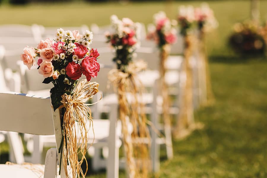 Fotografia dos detalhes de um casamento, com pequenos buquês de flores enfeitando as pontas das cadeiras dos convidados.